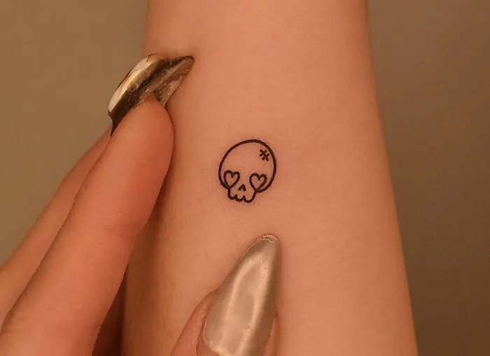 Minimal tattoo design of a small skull with heart eyes on an arm.