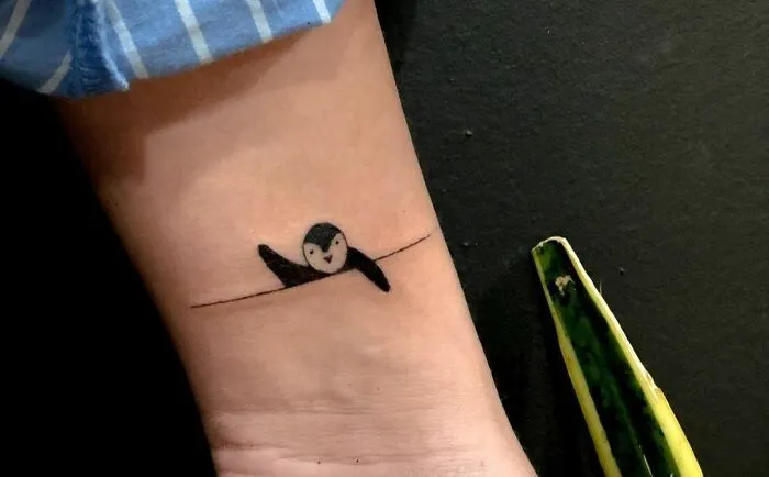 Minimal tattoo design featuring a small swimming penguin line art on an arm.