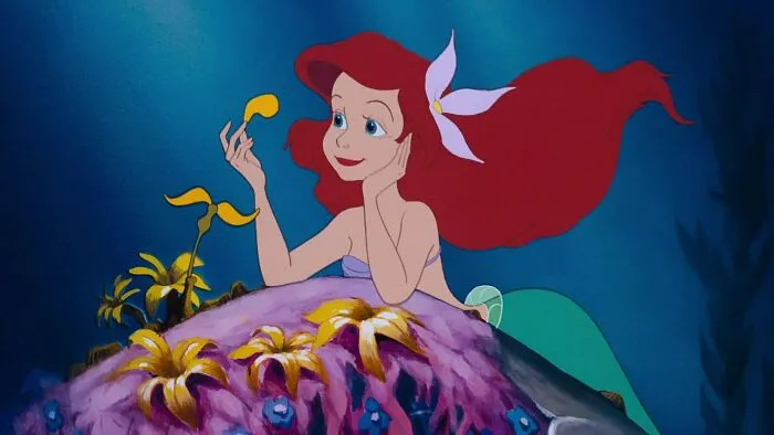 Animated female character with red hair and teal tail, sitting on a colorful ocean rock, smiling while holding a yellow fish.