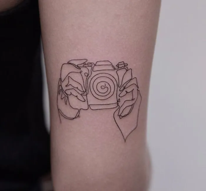 Fine-line single-line tattoo of hands holding a camera on an arm.