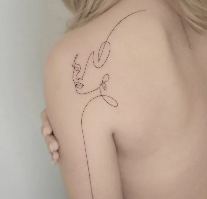 Fine-line tattoo of a face on a person's shoulder, showcasing single-line artistry.