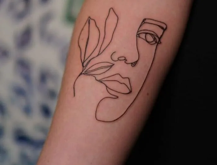 Fine-line tattoo of an abstract face with leaves on an arm.