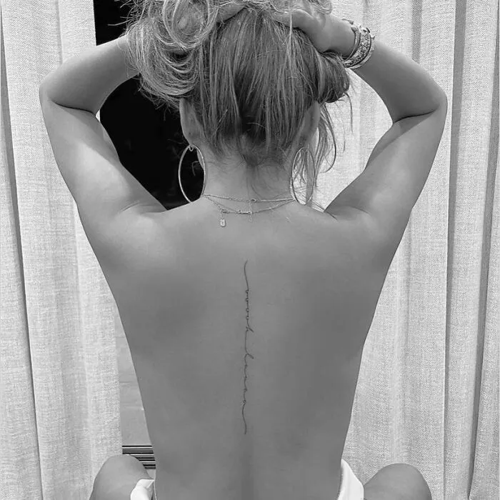 Celebrity tattoo on a woman's back, minimal script style, hair up, studio setting.