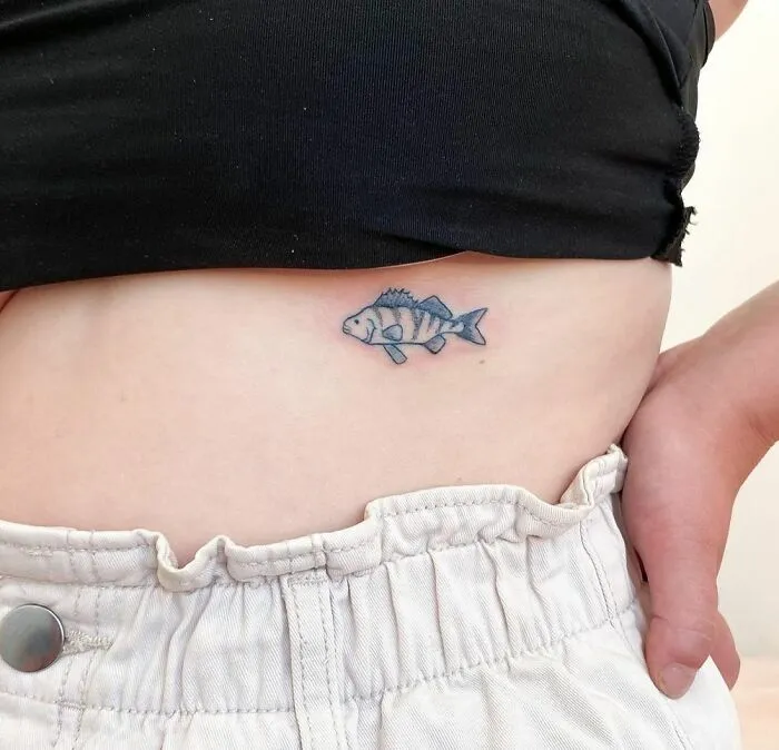 Minimalist tattoo of a fish on a person's side, above white pants, showcasing simple and aesthetically pleasing design.