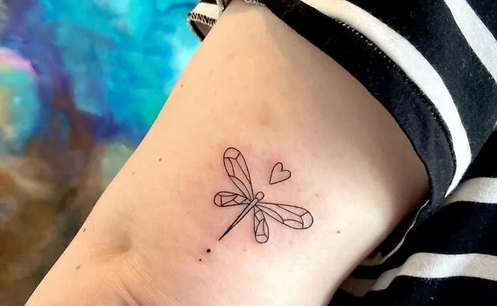 Minimalist dragonfly tattoo with a heart, inked on a person's arm, featuring clean lines in black ink.