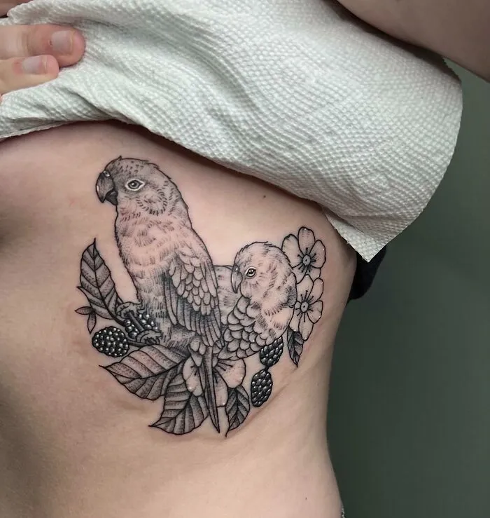 Pet tattoo featuring two detailed parrots with flowers and leaves on a person's rib cage.