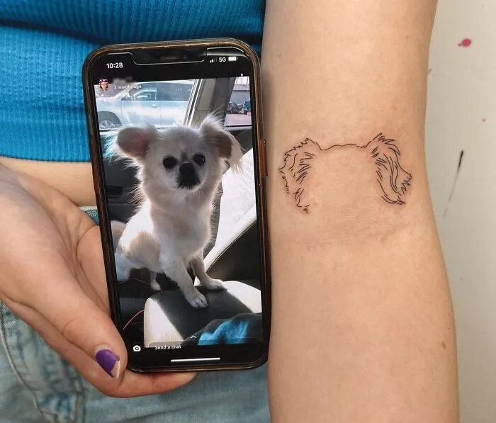 Pet tattoo of a dog's outline on an arm, with the dog's photo displayed on a smartphone nearby.