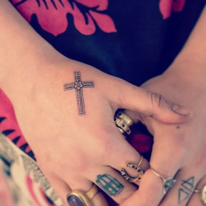 Close-up of a celebrity tattoo, a detailed cross on a hand with rings, against a floral fabric background.