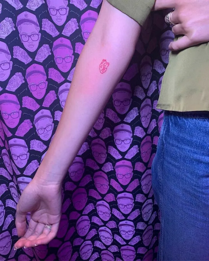Arm displaying a small red heart tattoo against a purple patterned background with celebrity faces.