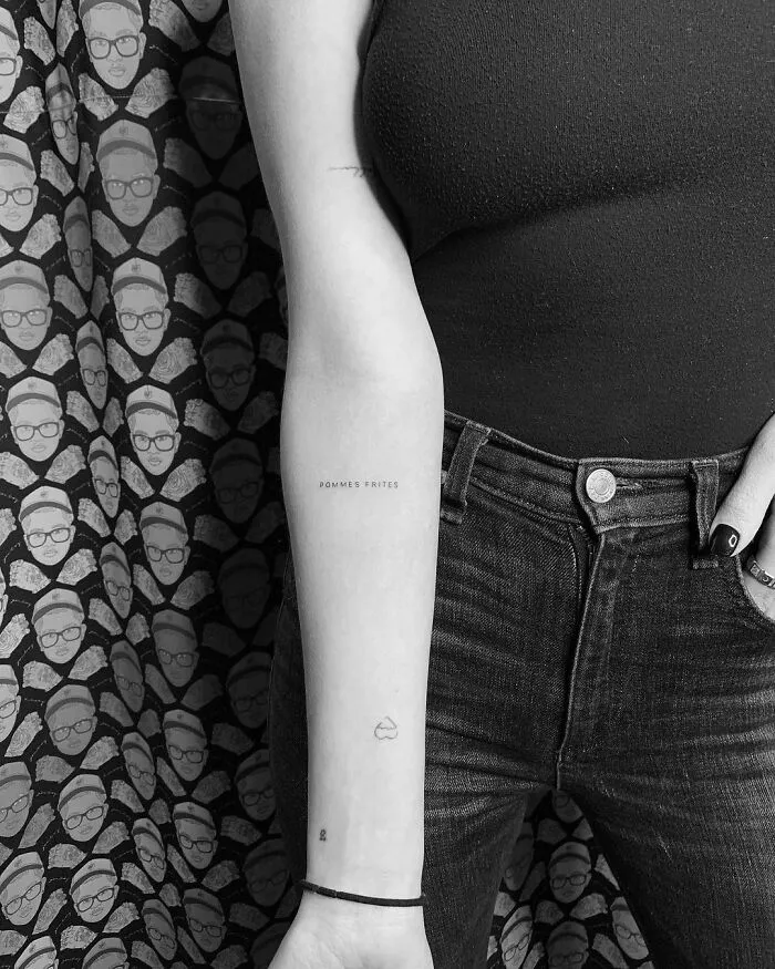 Close-up of forearm with celebrity tattoos, including text and small symbols, against a patterned background.