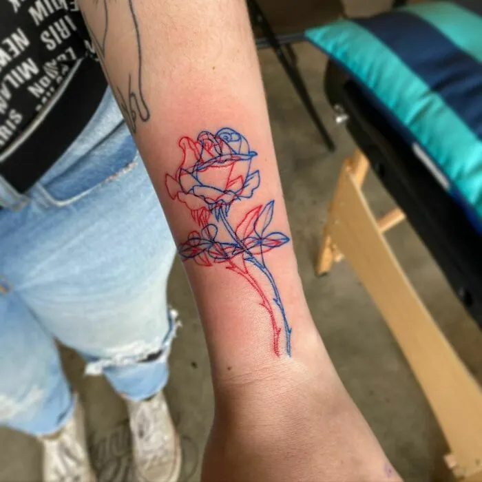 Optical illusion tattoo design of a rose in red and blue ink on an inner forearm.