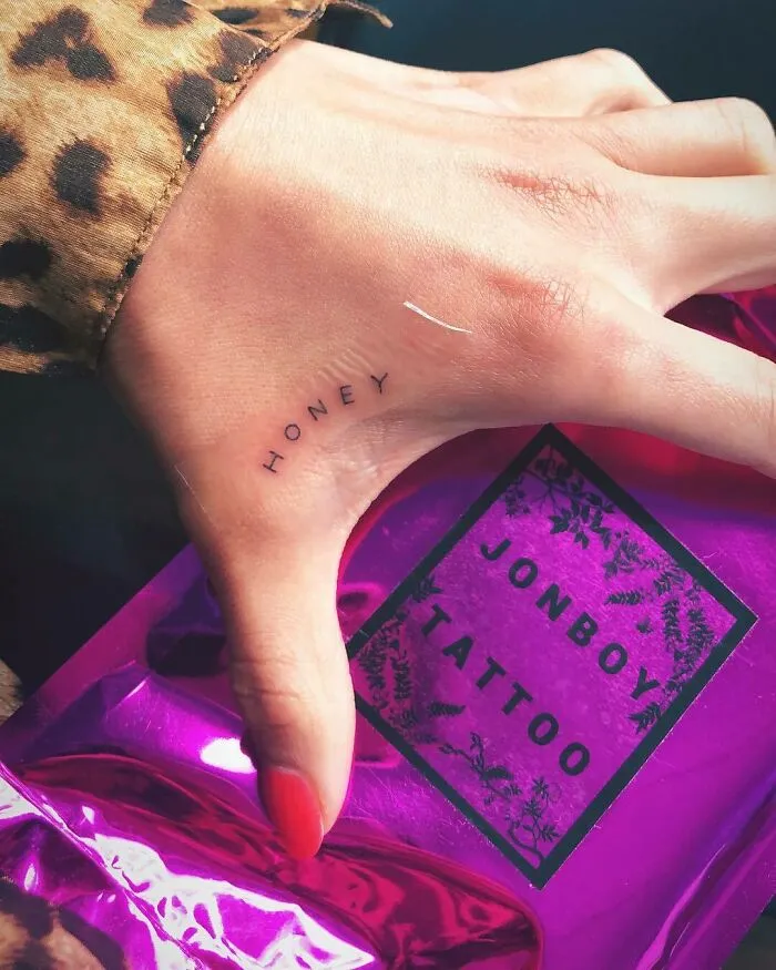 Close-up of a hand with "Honey" tattooed on the thumb, holding a pink Jonboy Tattoo bag.