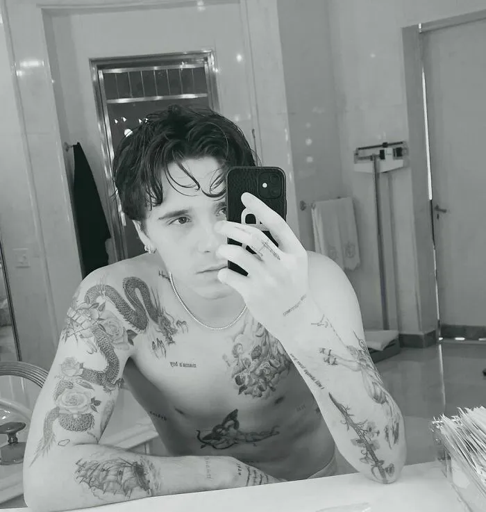Man with celebrity tattoos takes a mirror selfie in a bathroom.