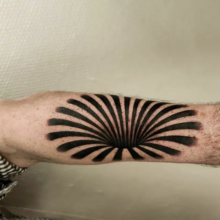 Optical illusion tattoo on an arm with black geometric design creating a 3D effect.