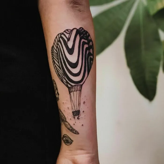 Optical illusion tattoo of a hot air balloon on a forearm, featuring bold, swirling patterns.