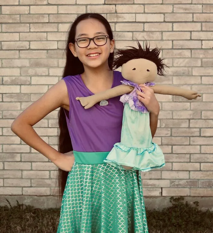 A smiling girl dressed as a mermaid, holding a custom doll that is a representation of herself.