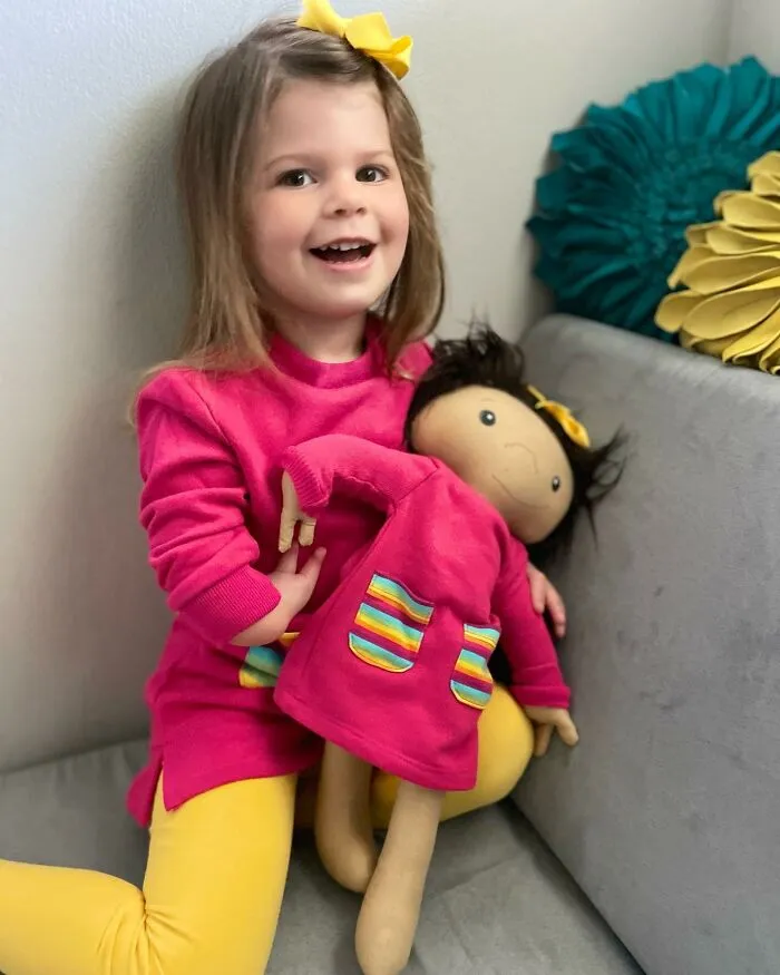 A happy young girl in a pink top and yellow leggings, embracing a custom doll wearing matching clothes, a representation of kids.