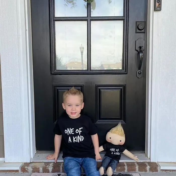 A boy wearing a 'One Of A Kind' shirt, sitting with a custom doll that looks like him, offering representation for kids.