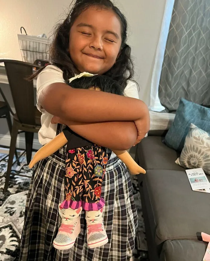 A smiling girl hugging her custom doll, both with dark hair, celebrating diverse doll representation.