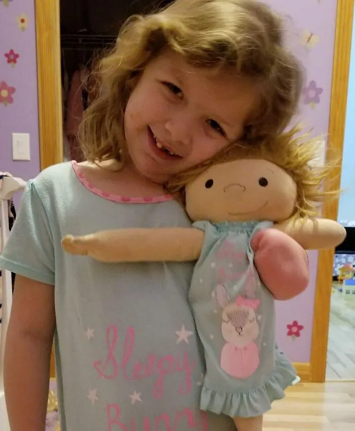 A girl with curly hair in a light blue shirt smiles, holding her custom doll, which is a representation of herself.