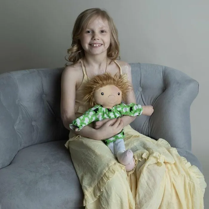 A smiling girl with light hair, sitting on a grey couch and holding a custom doll that represents her.