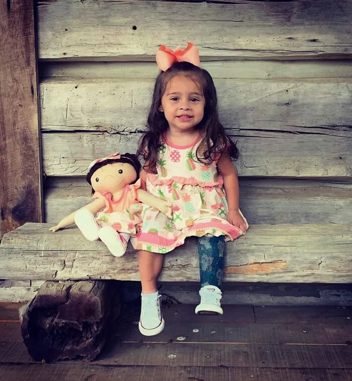 A little girl with a prosthetic leg sits next to her custom doll representation, both smiling on a bench.