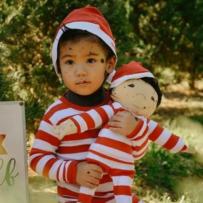 A young boy in a red and white striped outfit and hat, holding a custom doll that represents him.