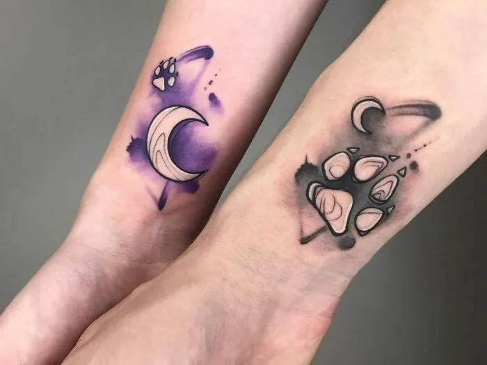 Watercolor tattoos of crescent moons and paw prints on forearms, blending vibrant purple and black hues.