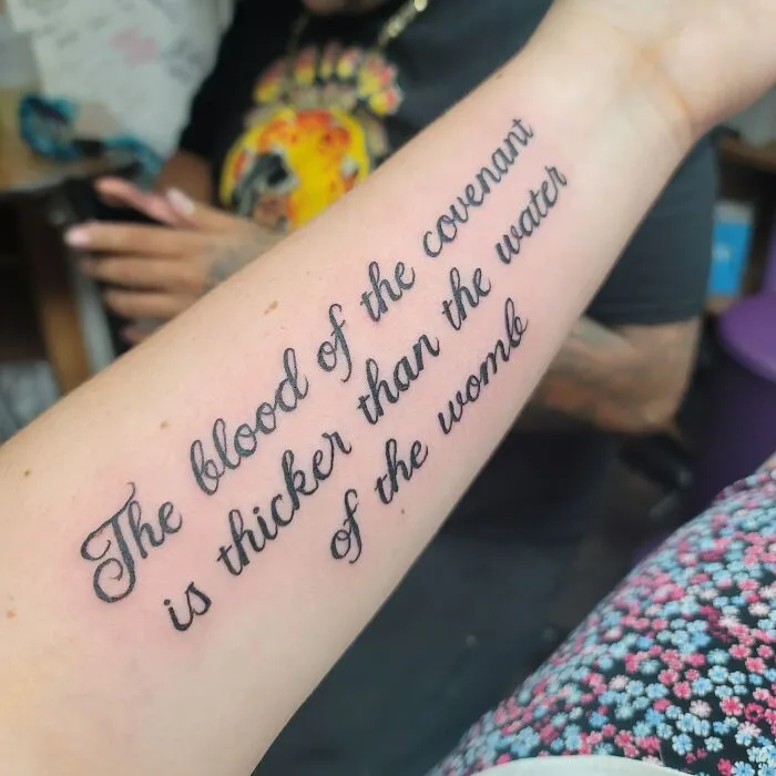 Word tattoo on forearm reading "The blood of the covenant is thicker than the water of the womb" in black script.