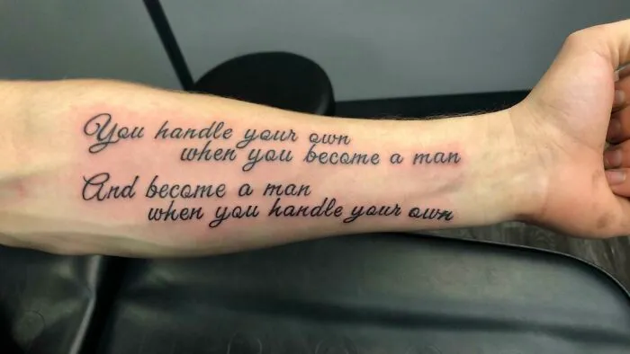 Arm with a word tattoo in elegant script, featuring an inspirational quote about maturity and self-reliance.