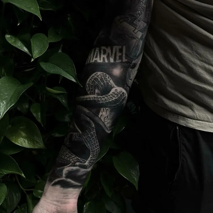 Detailed Marvel tattoo sleeve with iconic elements on a person's arm, surrounded by green leaves.