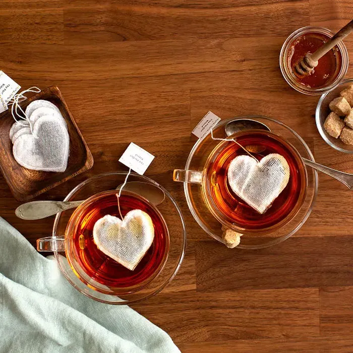 Heart-shaped tea bags in cups for Valentine's Day gifts, with honey and sugar cubes on a wooden table.