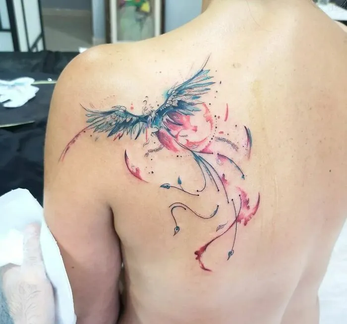 Watercolor tattoo of a bird on a person's shoulder, featuring vibrant blue and red hues.