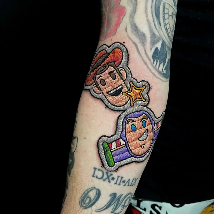 Embroidered-style tattoo by Eduardo Lozano featuring cartoon characters Woody and Buzz on a person's forearm.