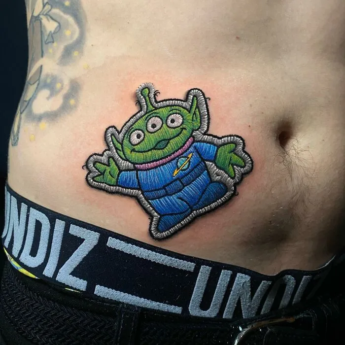 Tattoo by Eduardo Lozano featuring a colorful three-eyed alien on a person's abdomen.
