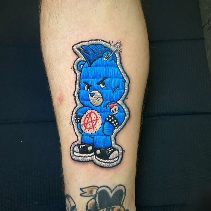 Embroidered-style tattoo of a blue bear with punk details on a person's leg, showcasing a new dimension in tattooing.