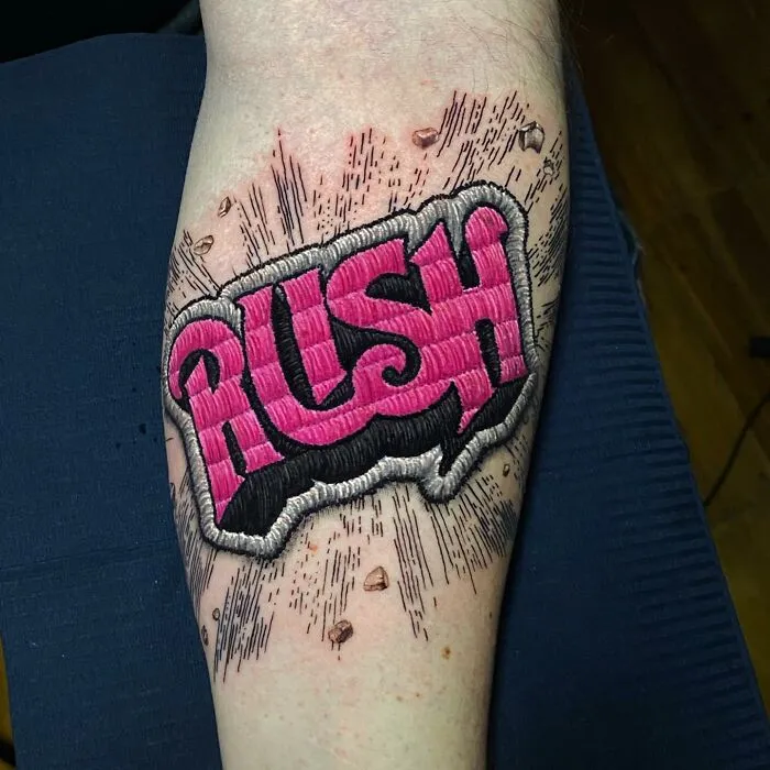 Tattoo by Eduardo Lozano showcasing "RUSH" in 3D embroidery style on a forearm.