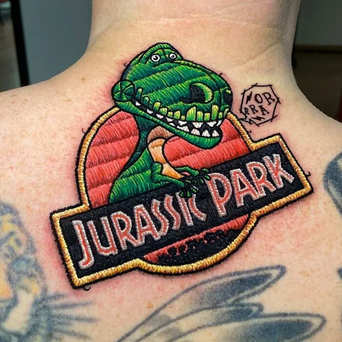 Tattoo of Jurassic Park logo with embroidered style by Eduardo Lozano on the back of a person's neck.