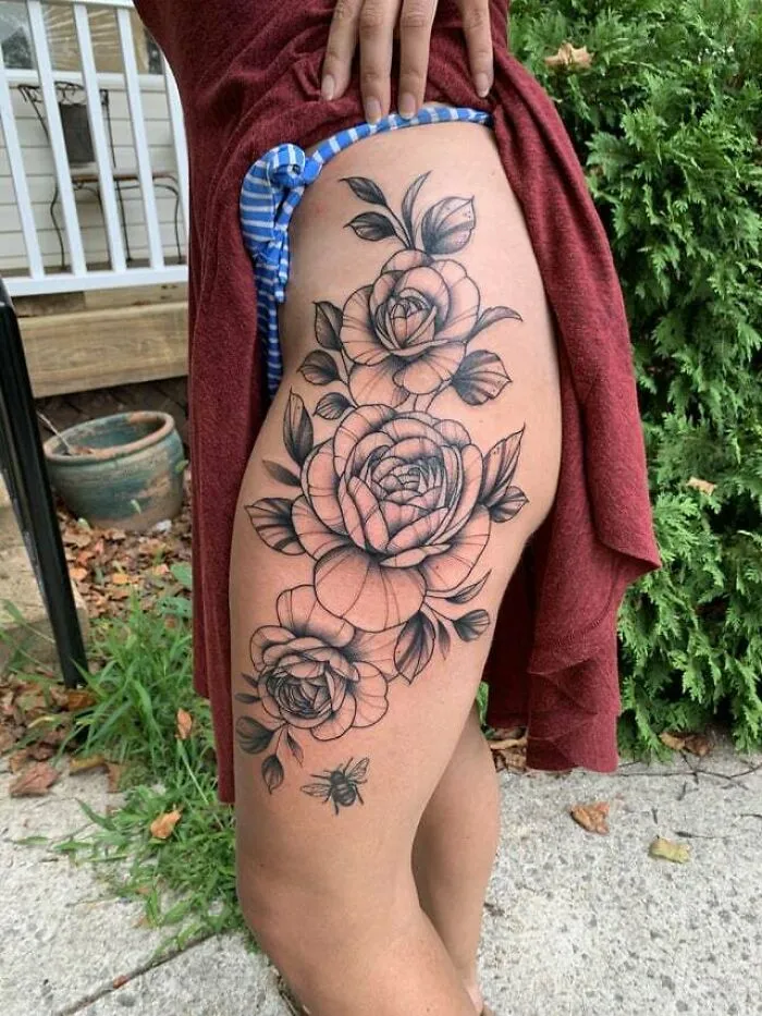 Elegant flower tattoo on a person's thigh, showcasing intricate rose designs with leaves and a small bee.