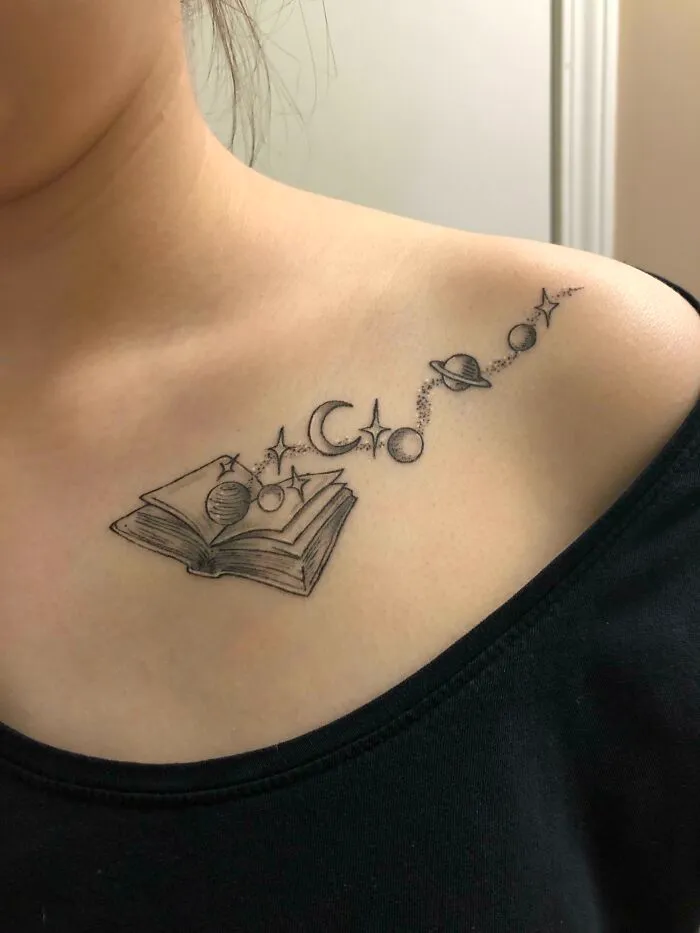 Space tattoo on a shoulder featuring open book, planets, stars, and a crescent, symbolizing outer space themes.