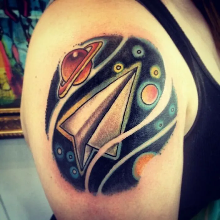 Space tattoo featuring a paper airplane surrounded by planets and cosmic elements on an arm.
