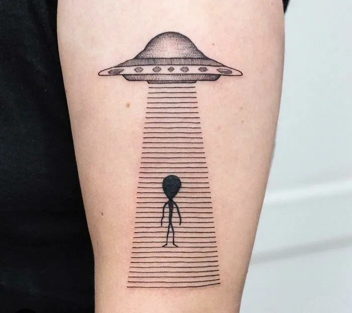 Alien and UFO space tattoo on forearm, featuring a spacecraft beaming up a stick-figure alien illustration.