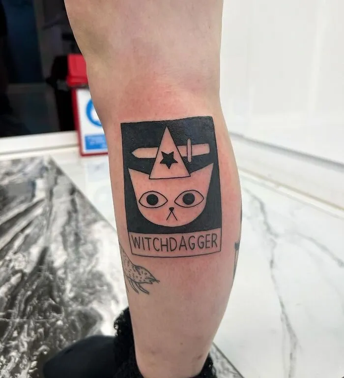 Calf tattoo featuring a cat with a hat and dagger, labeled "Witchdagger," showcasing cool tattoo art.