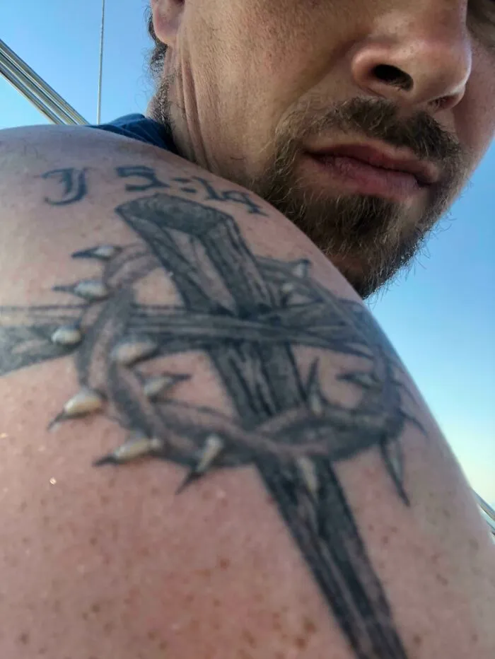 Man with a tattoo of a cross and thorn crown on shoulder, perfectly matching the moment.