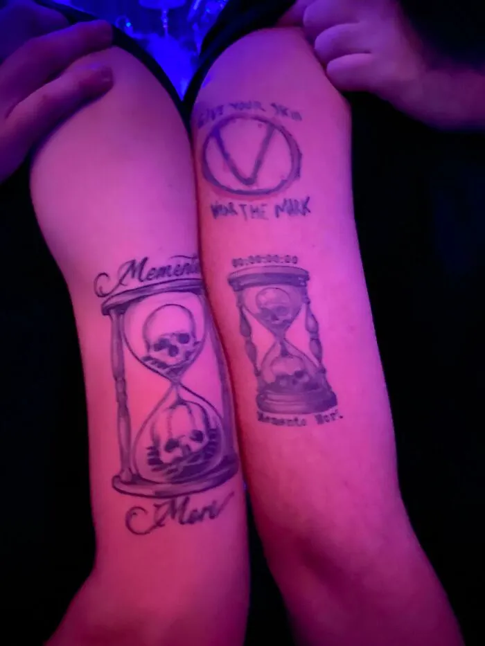 Matching tattoos with skulls and hourglasses on arms, perfectly aligned in a low-light setting.