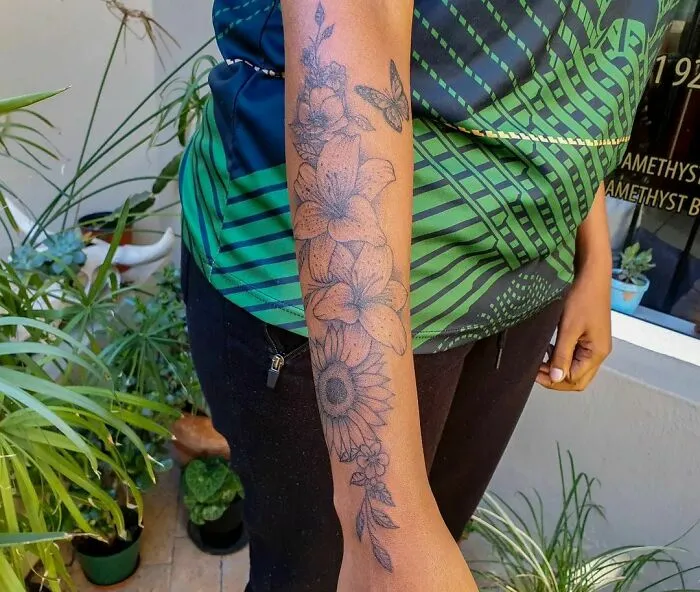 Arm adorned with detailed flower tattoo, showcasing elegance and beauty among lush green plants.