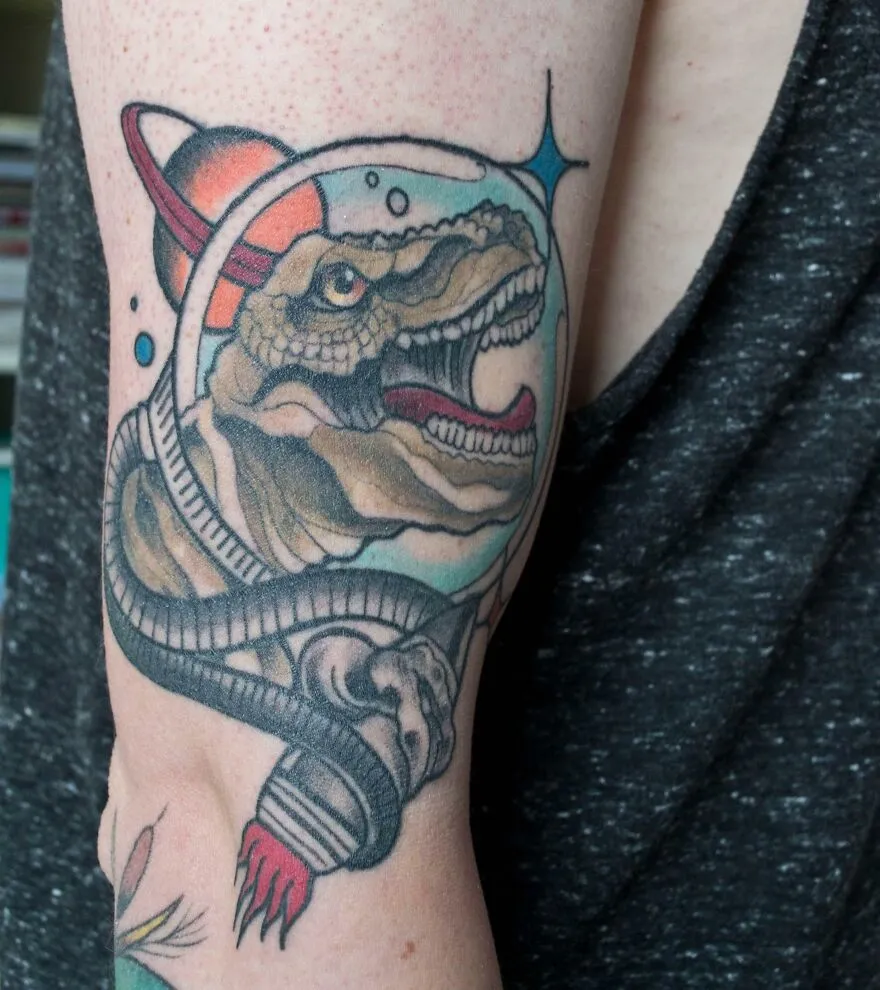 Space tattoo of a dinosaur in an astronaut helmet, featuring stars and a planet, on an arm.