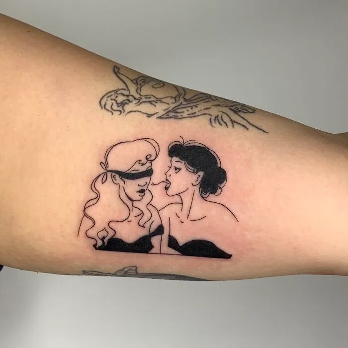Illustrative tattoo of two women, one blindfolded, on an arm.