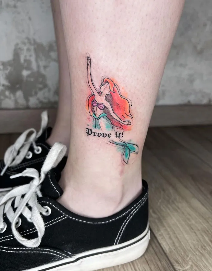 Watercolor tattoo of a mermaid with "Prove it!" text on a person's leg, near black sneakers.