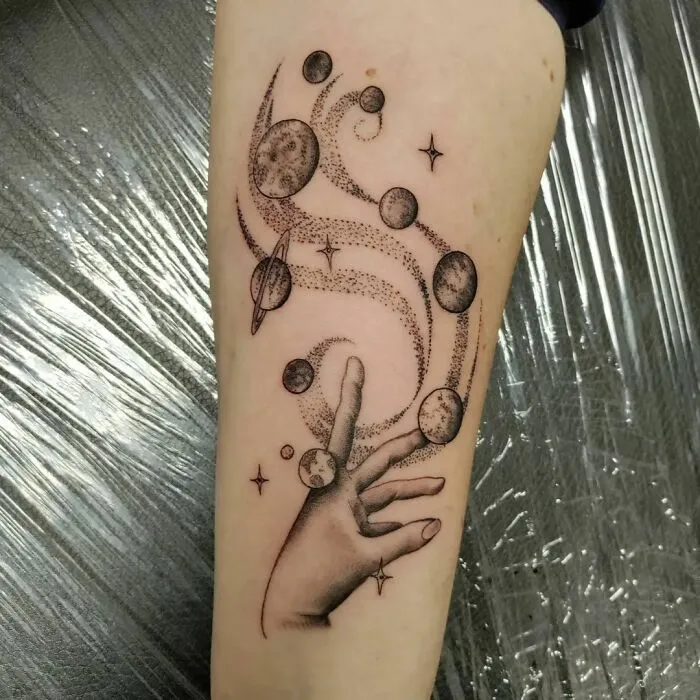 Space tattoo featuring a hand reaching for planets and stars in a cosmic design on forearm.